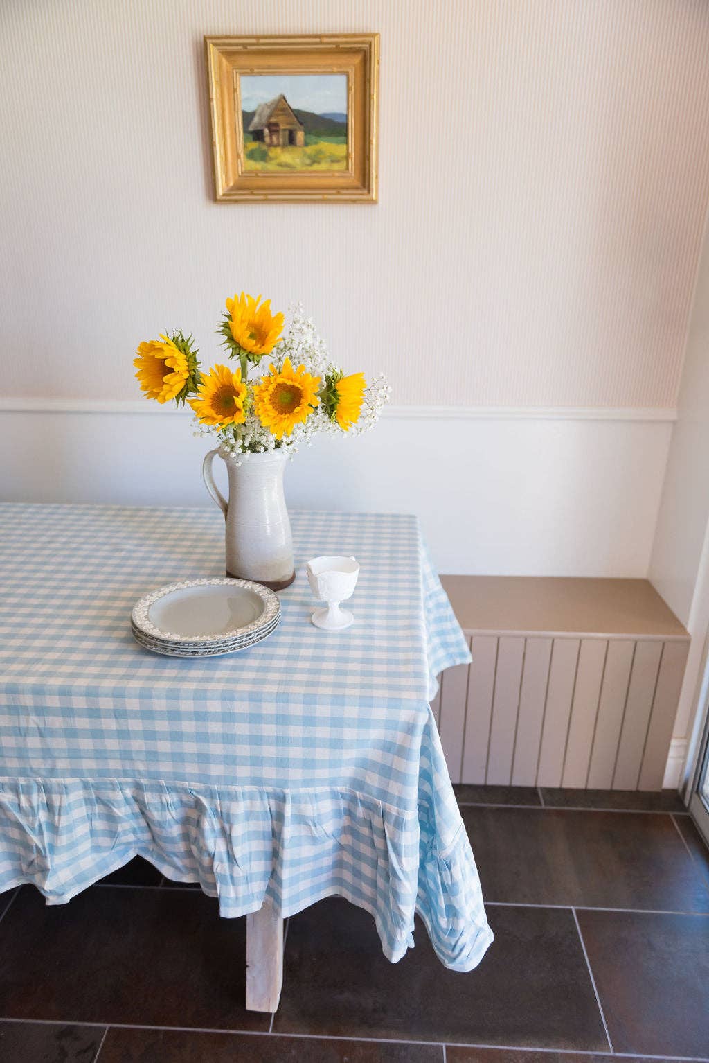 Table with a blue checkered tablecloth, sunflowers in a vase, and a painting on the wall.