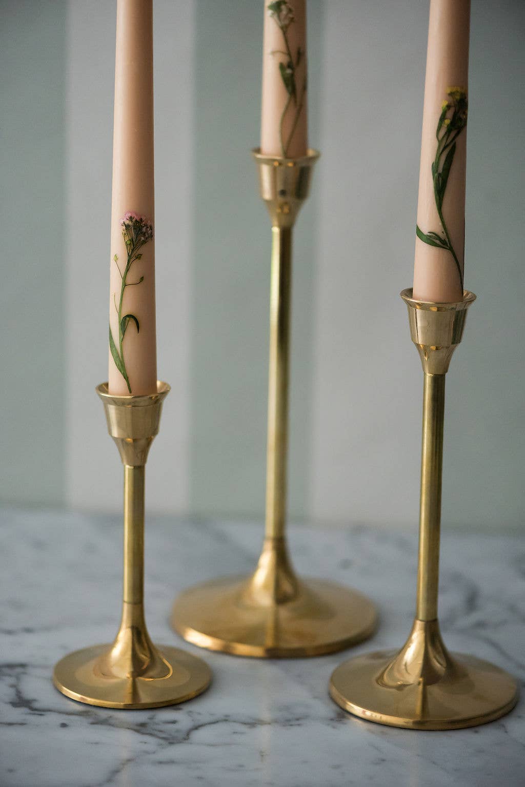 Three gold candle holders with candles featuring floral designs on a marble surface.