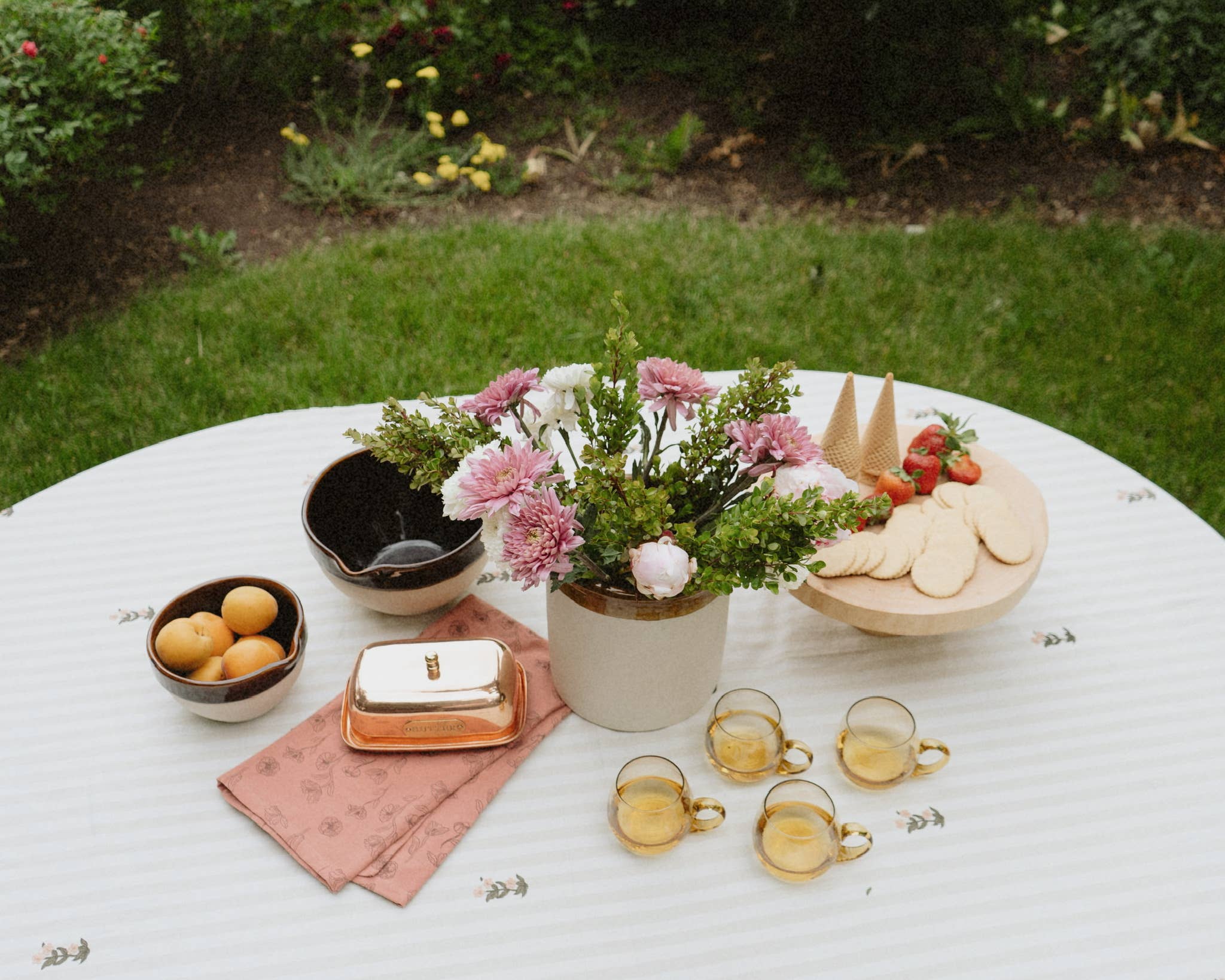 Outdoor setting with a table set for a meal, including flowers, fruit, and cutlery.