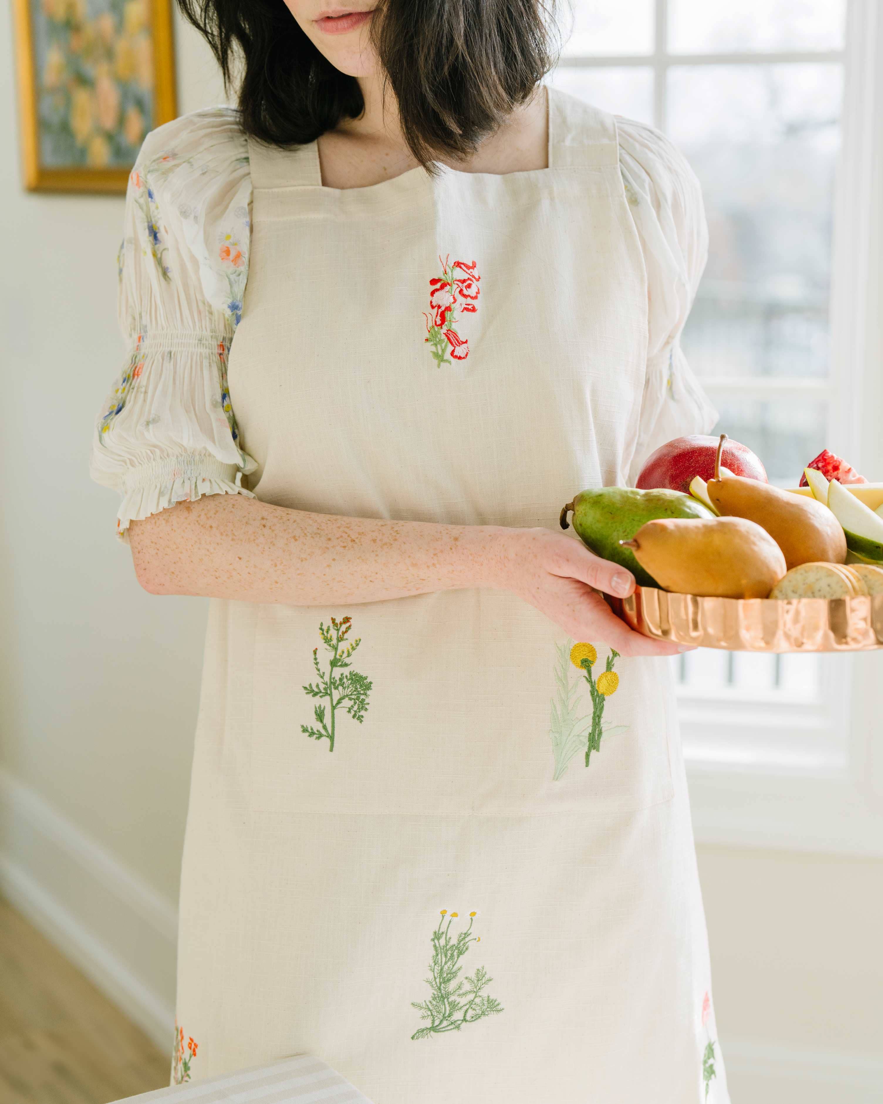 Person wearing a beige apron with floral embroidery, holding a basket of fruits.