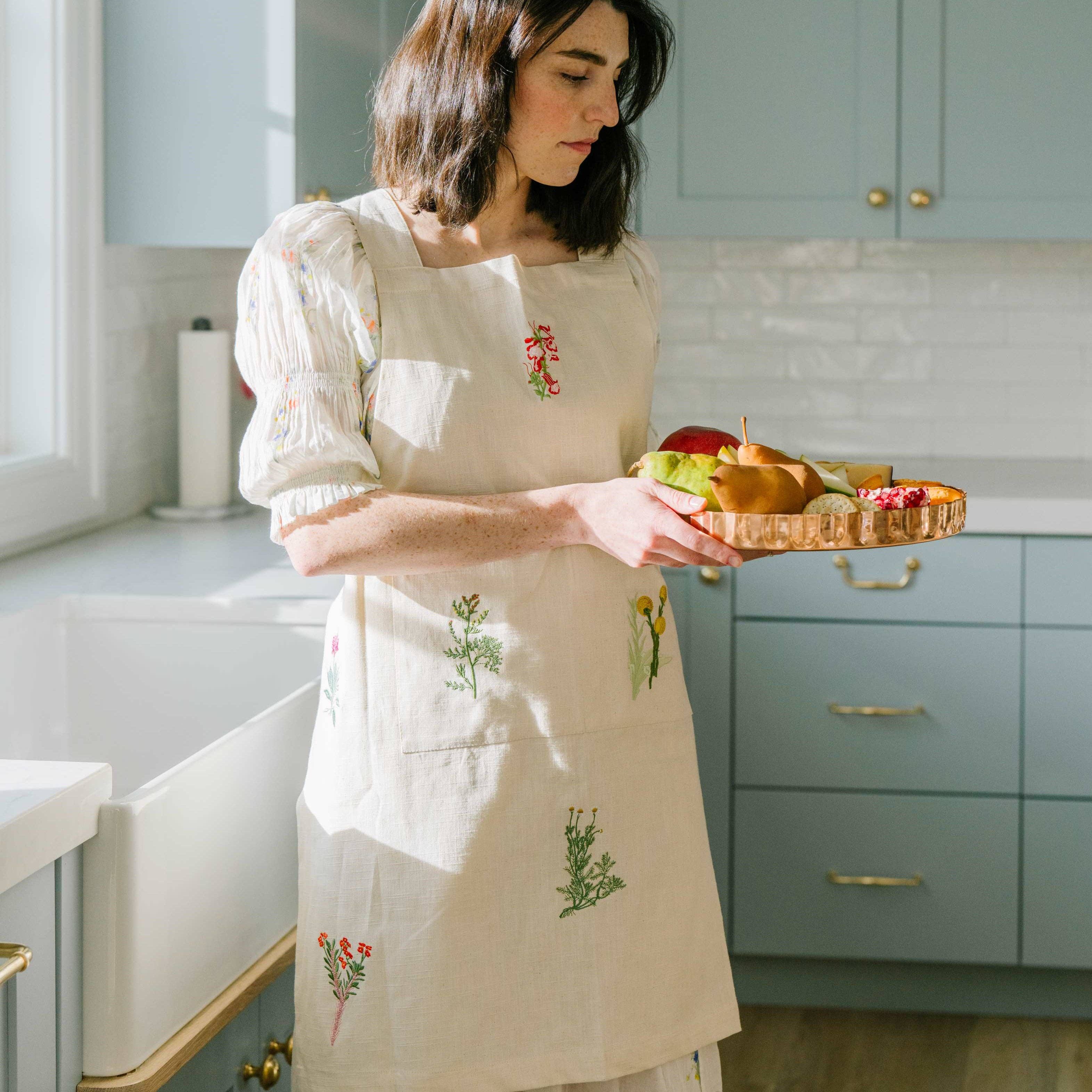 Woman in a kitchen holding a basket of fruits and vegetables wearing an apron with floral embroidery.