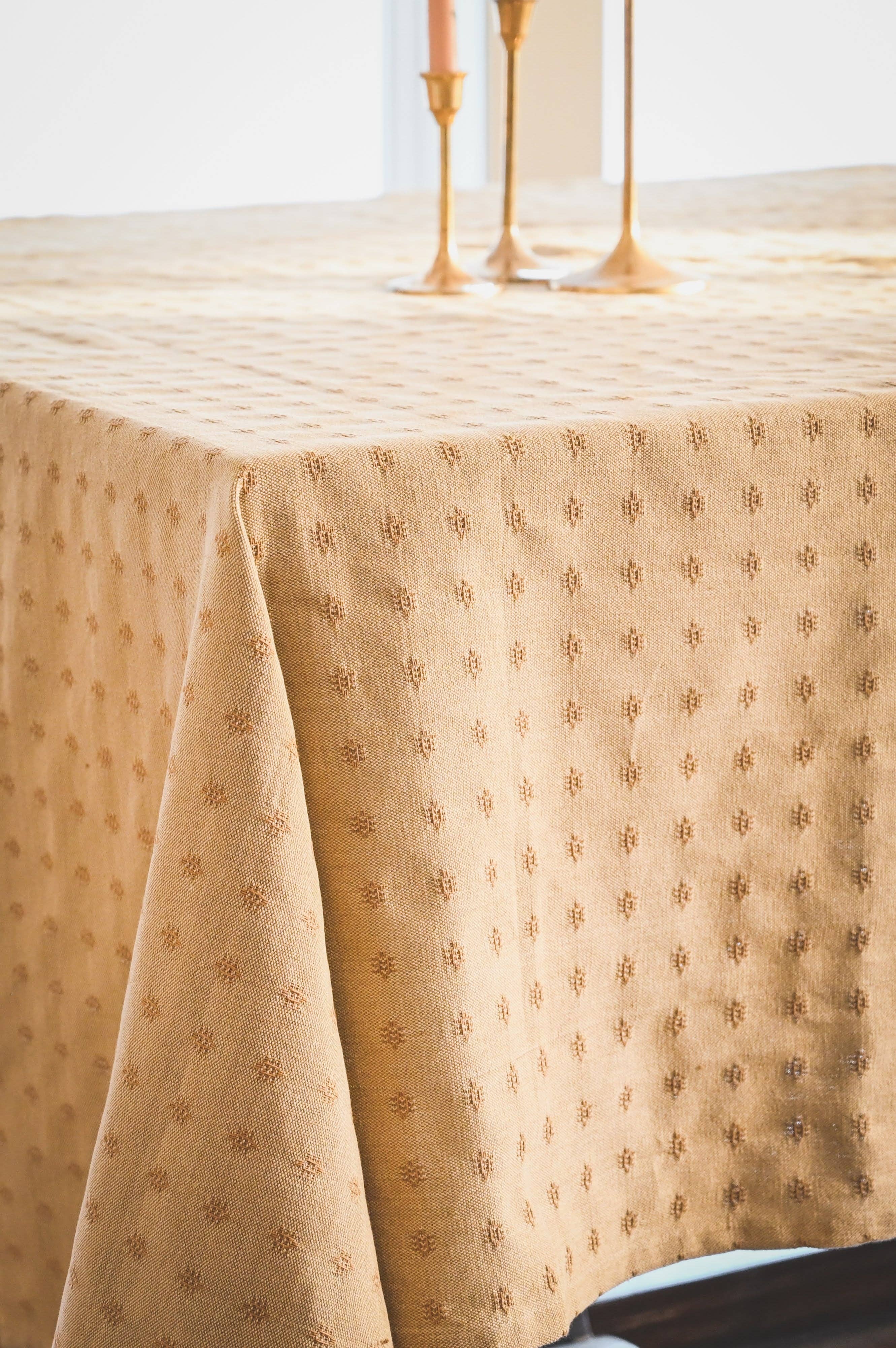 Tan tablecloth with subtle pattern on a table, with candles in the background.