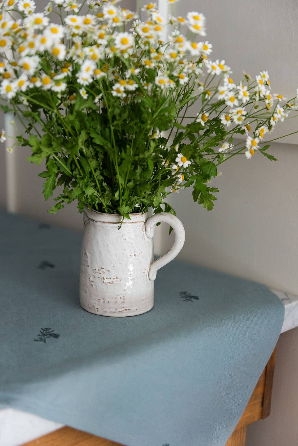 White ceramic pitcher with greenery and white flowers on a green-blue tablecloth.
