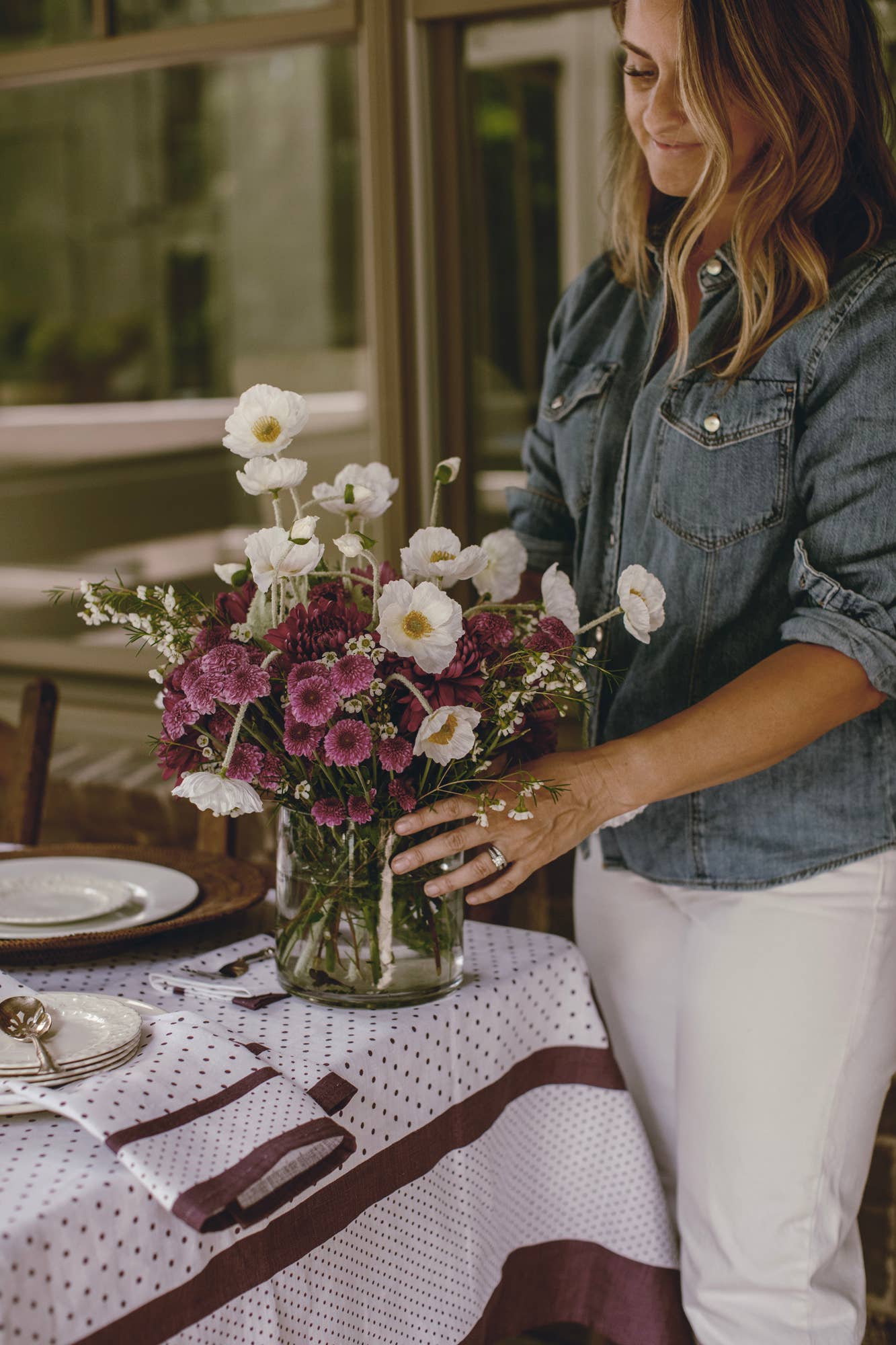 Woman arranging flowers in a vase on a table with a floral tablecloth.