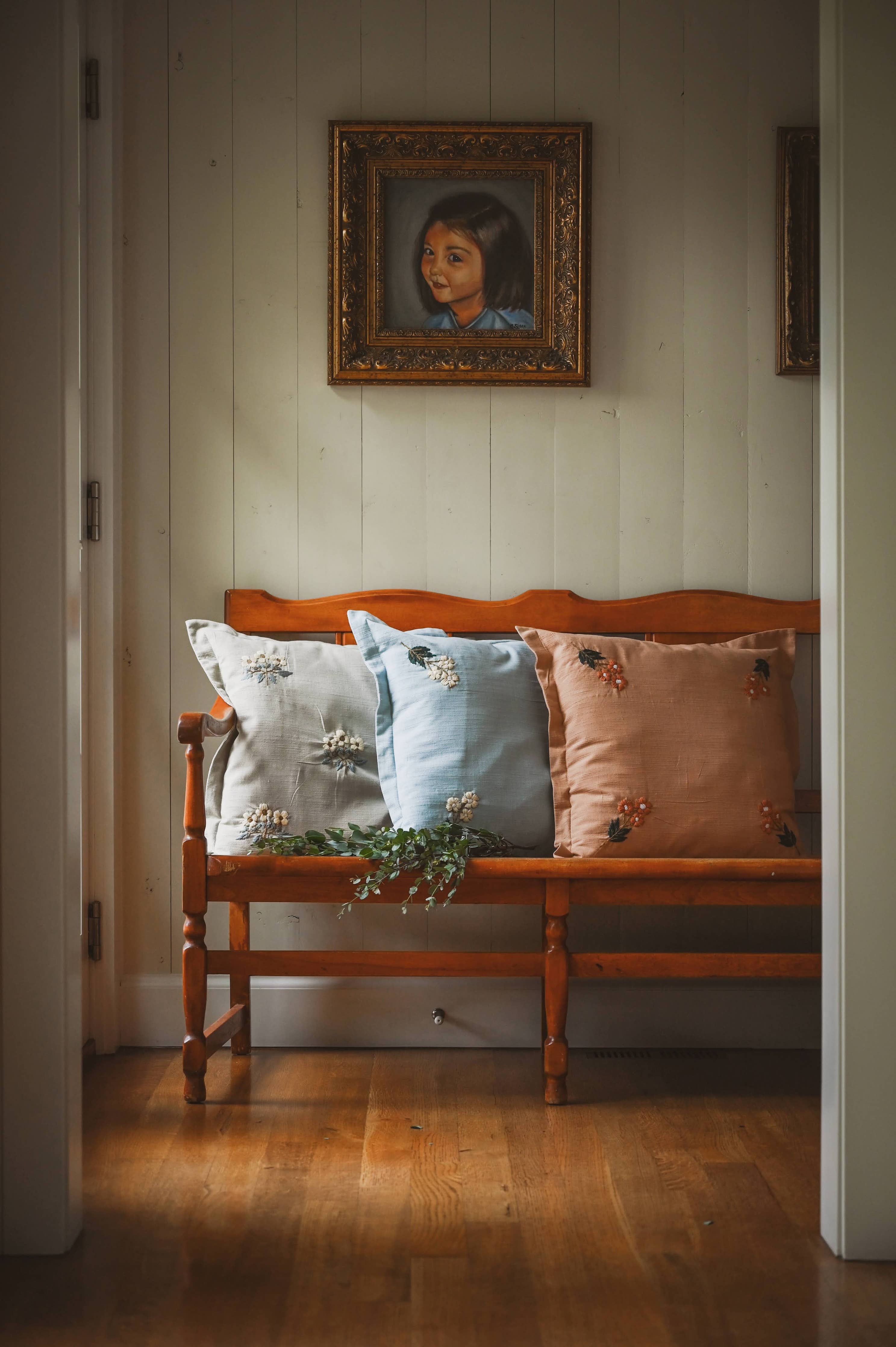 Wooden bench with embroidered pillows against a white paneled wall with a framed portrait.