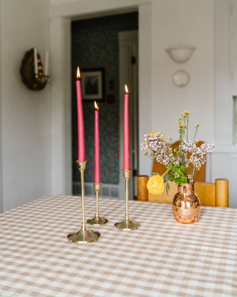 Pink candles in brass candleholders on a gingham tablecloth with a vase of flowers.