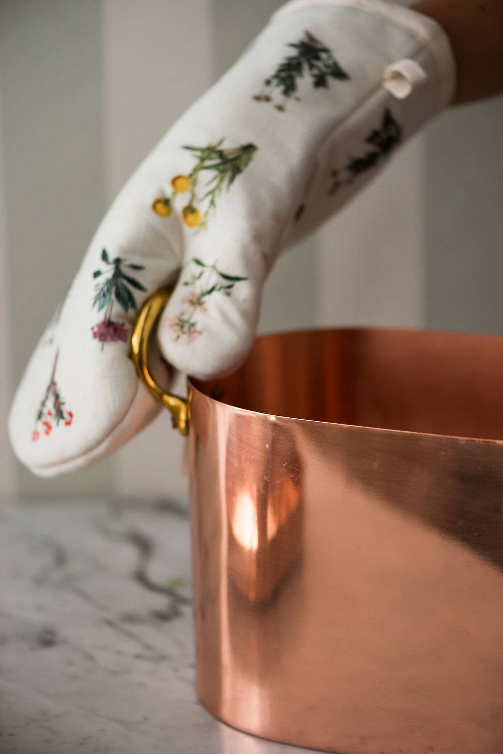 White oven mitt with floral patterns holding a copper pot.