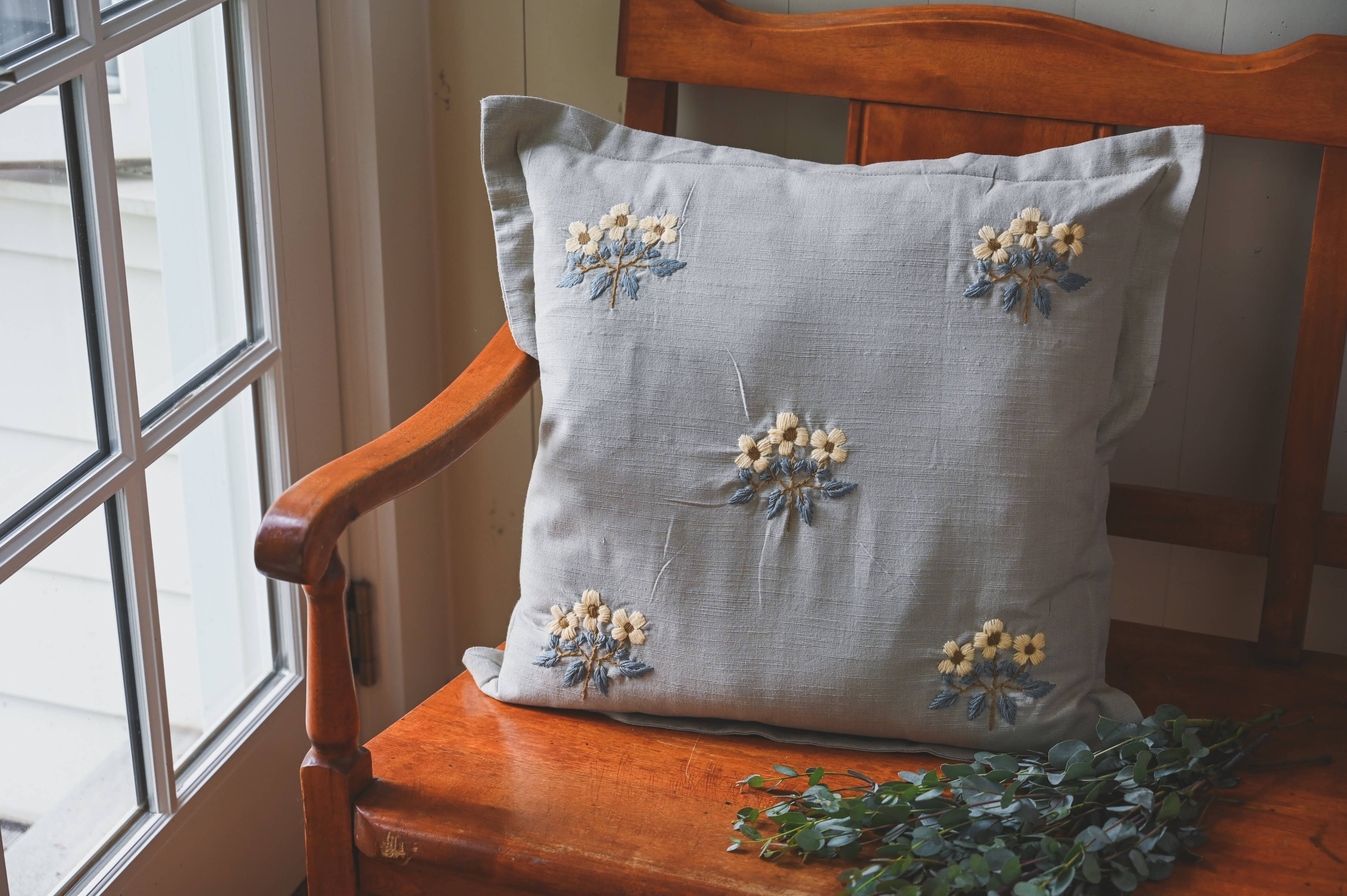 Decorative pillow with floral embroidery on a wooden chair by a window.