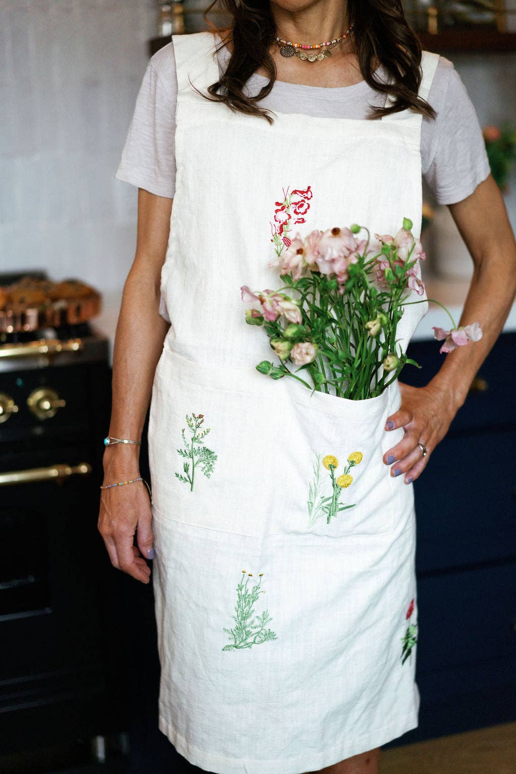 Person wearing a white apron with floral designs holding a bouquet of flowers in a kitchen.