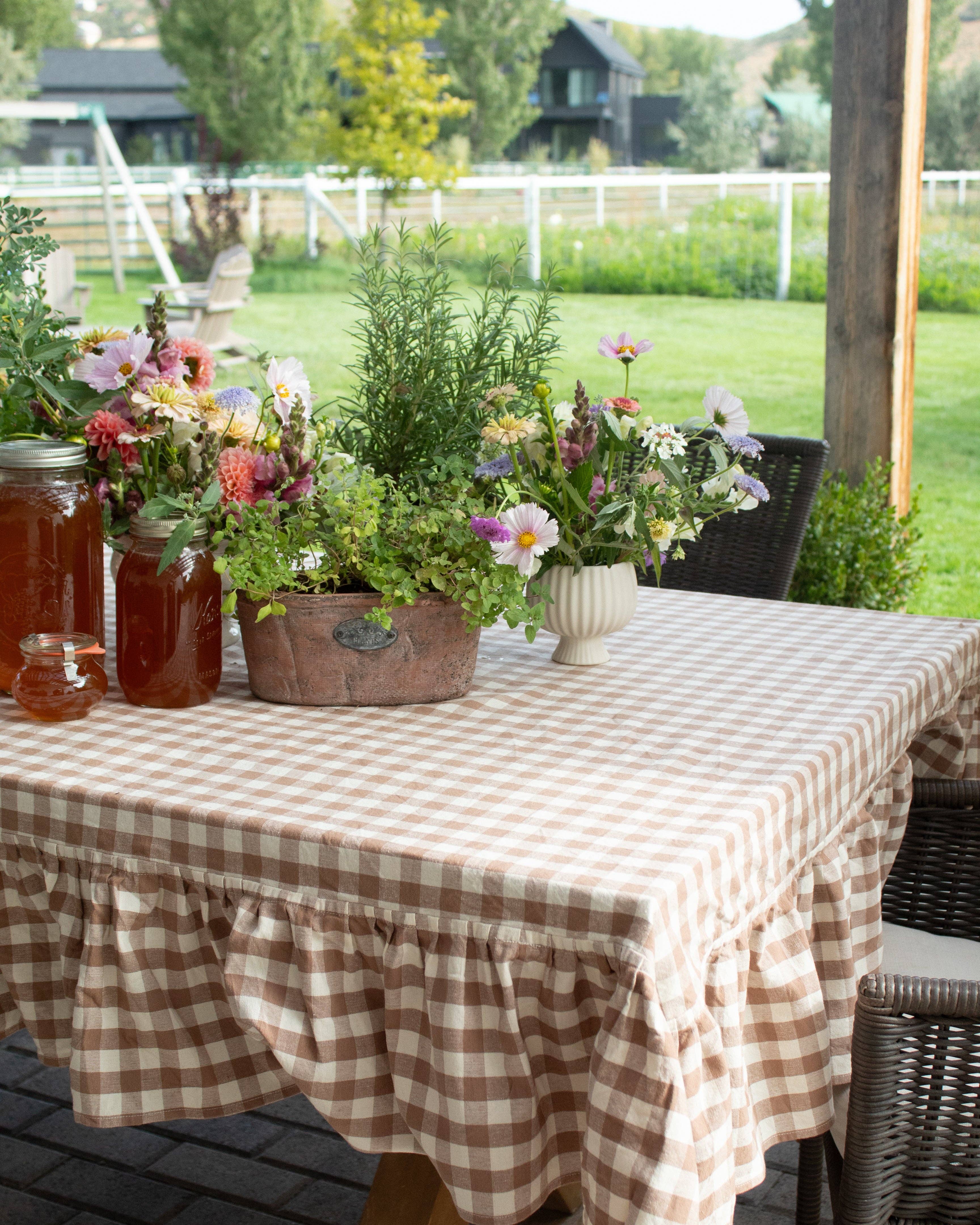 Outdoor setting with a table covered by a gingham ablecloth, jars of pickles, and floral arrangements.